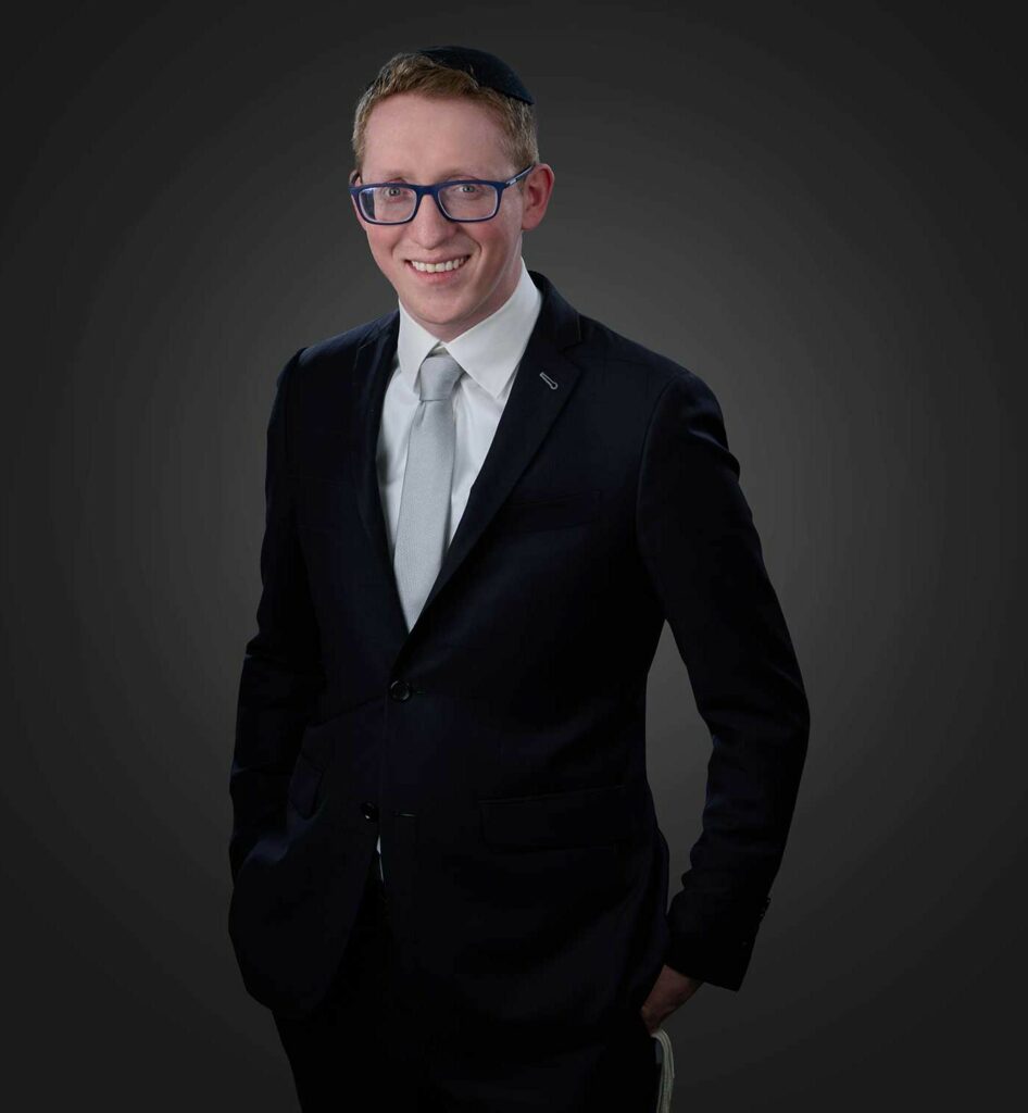 Smiling man wearing a dark suit with a light gray tie, blue glasses, and a black kippah, standing against a dark gray background with hands in pockets.