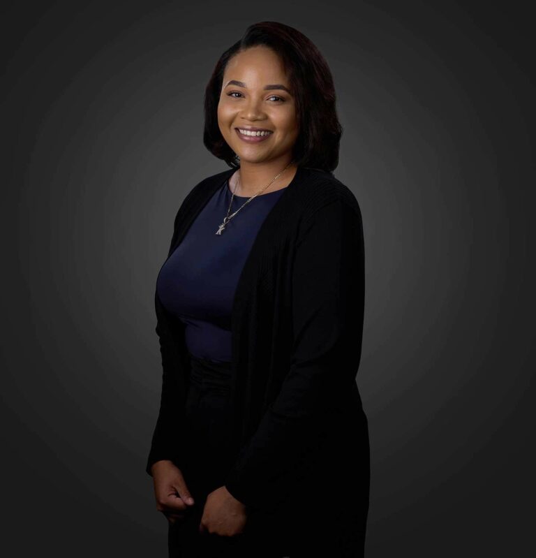 Portrait of a smiling woman with shoulder-length dark hair wearing a dark blue top, black cardigan, and necklace, standing against a dark gray background.