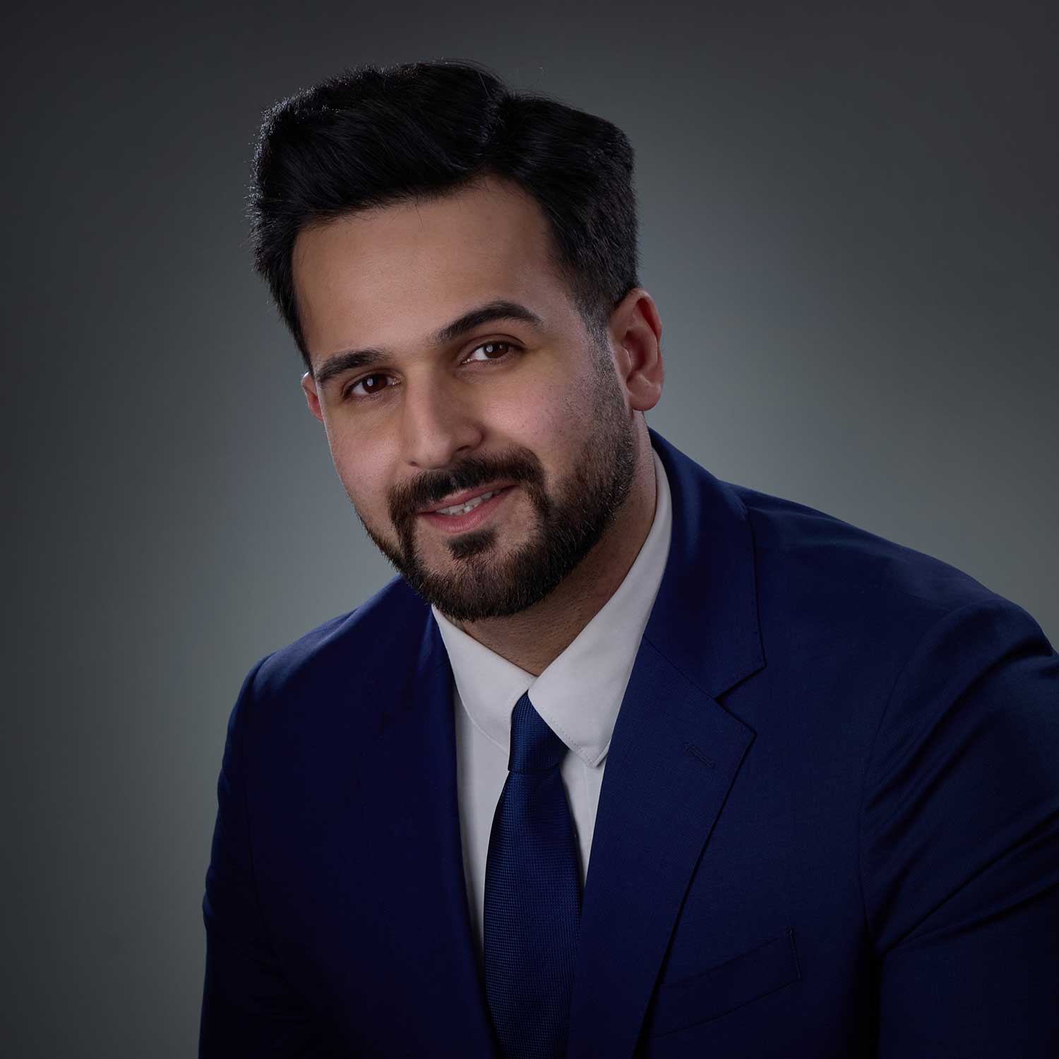Smiling man with dark hair and beard wearing a dark blue suit, white shirt, and dark blue tie against a gradient gray background.