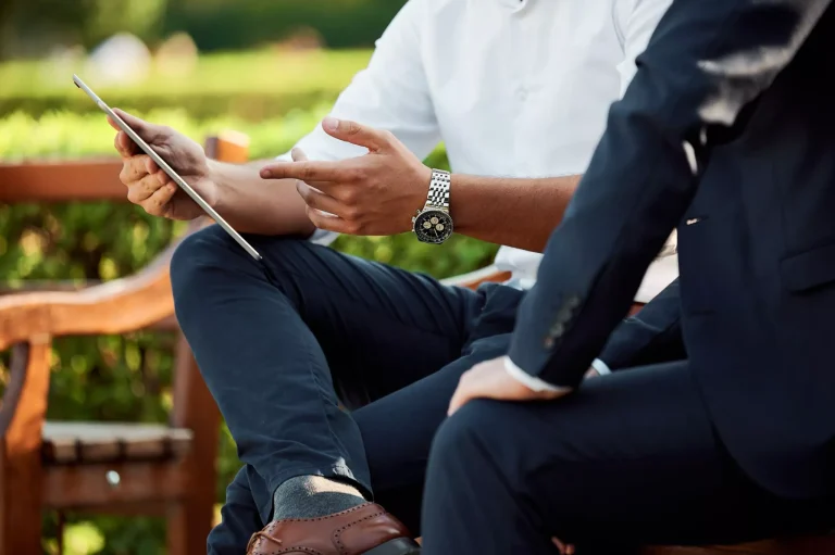 Two professionals, one in a white shirt and the other in a suit, sitting on a wooden bench outdoors discussing information on a tablet, symbolizing consultation with Nussbaum family lawyers in Toronto.