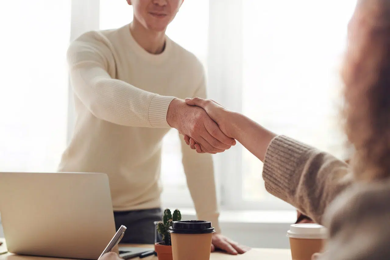 Two people shaking hands across a desk with a laptop and coffee cups, symbolizing a professional agreement, representing Nussbaum family lawyers in Toronto providing client support.
