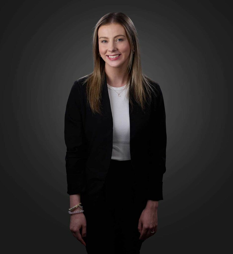 Smiling young woman with long straight hair wearing a black blazer over a white top standing against a dark gray background