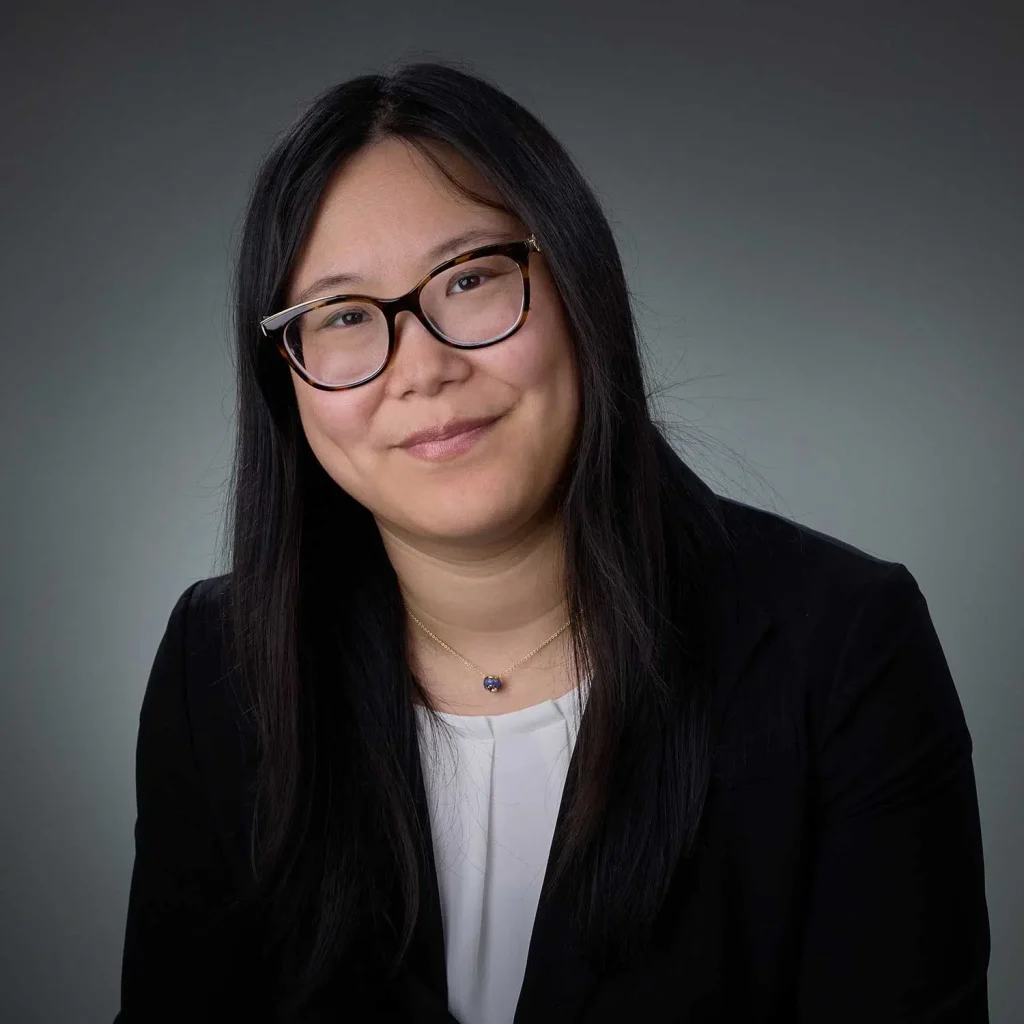 Portrait of a woman with long black hair and tortoiseshell glasses, wearing a black blazer over a white top, smiling gently against a gray background.
