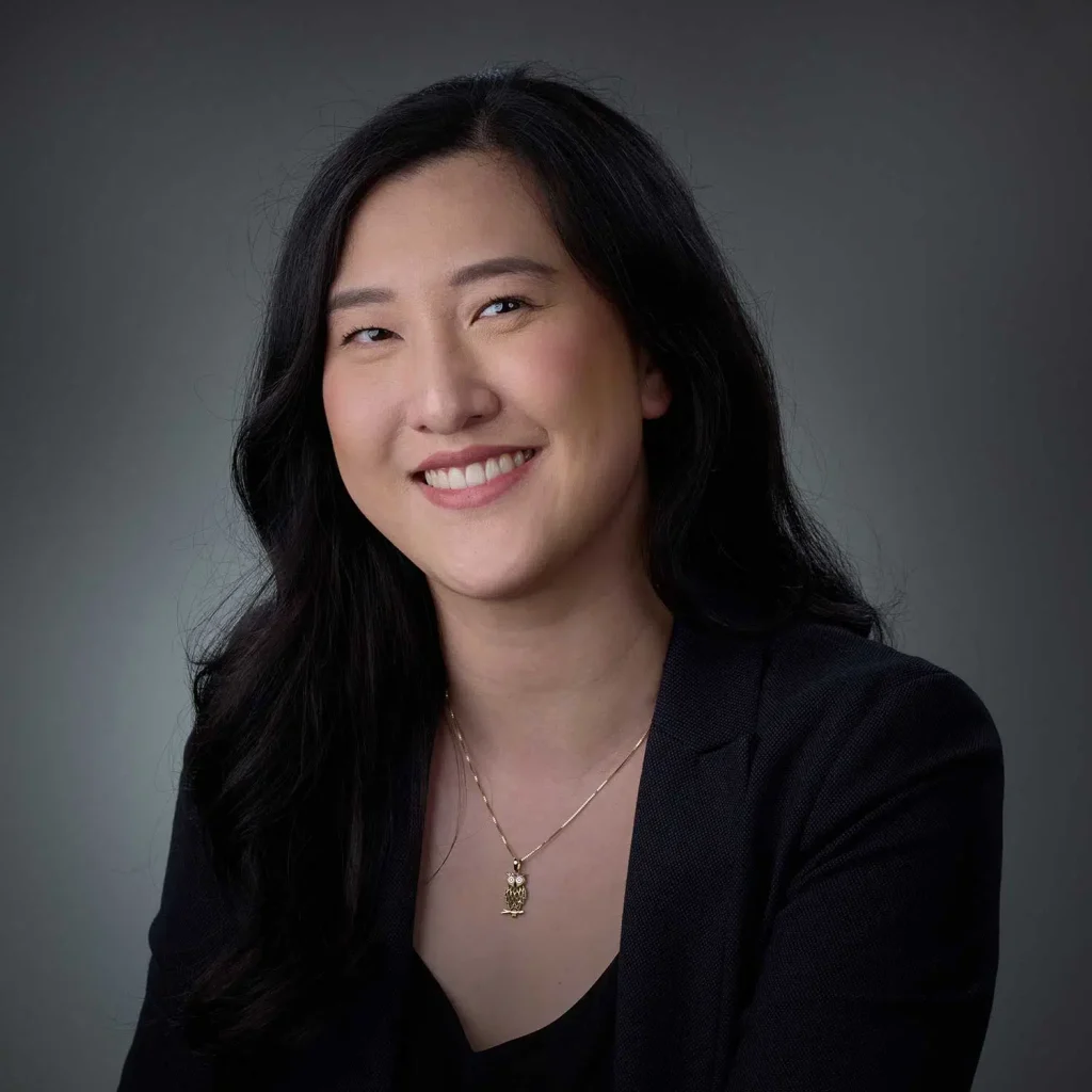 Portrait of a smiling woman with long black hair wearing a black blazer and a gold owl pendant necklace against a grey background