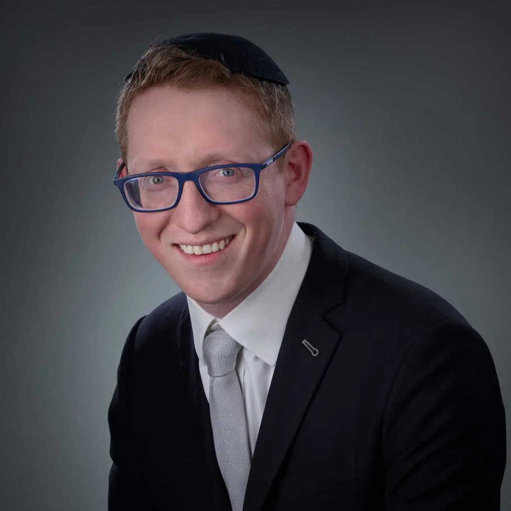 Smiling man wearing blue glasses, a black kippah, a dark suit, white shirt, and light gray tie against a gradient gray background.