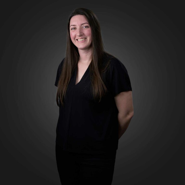 Smiling woman with long straight brown hair wearing a black short-sleeve V-neck top and black pants standing against a dark gray background with her hands behind her back
