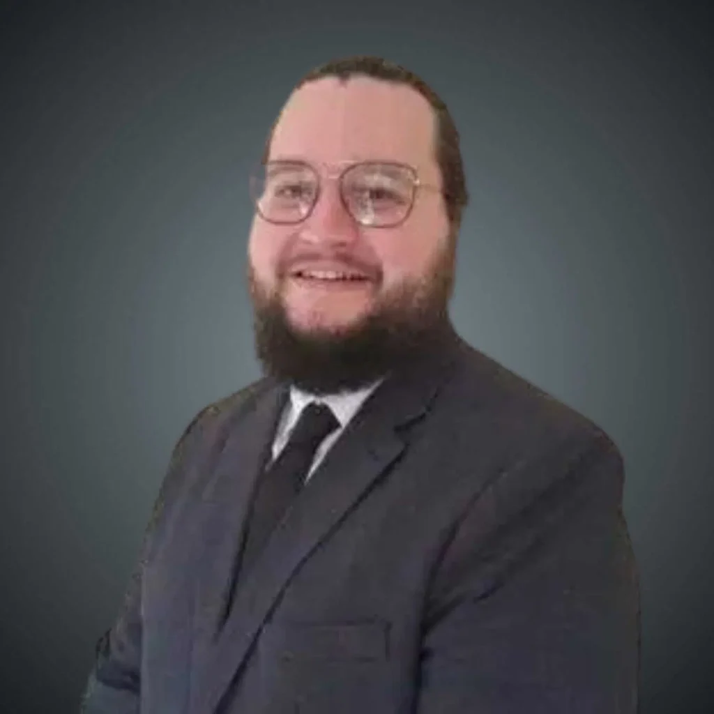 Smiling man with a beard and glasses wearing a dark suit, white shirt, and black tie against a gray background