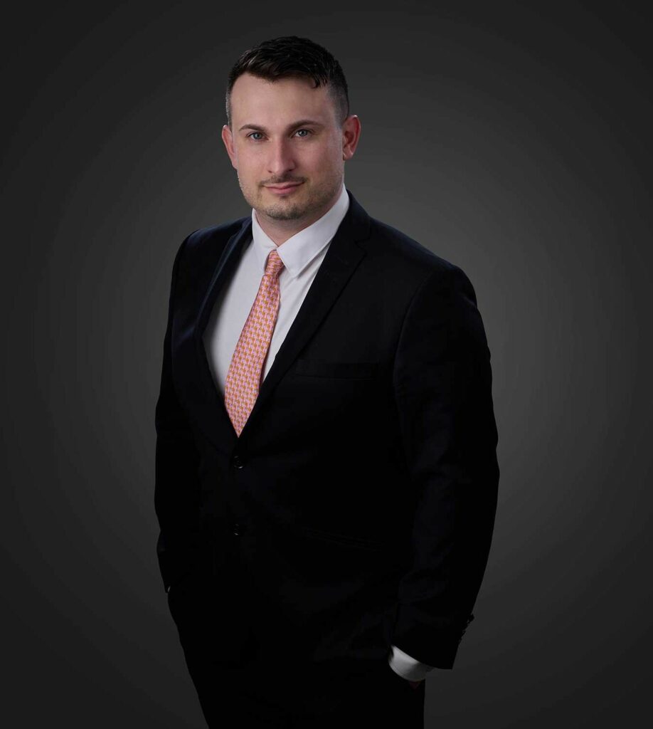 Man in a black suit, white dress shirt, and patterned orange tie standing against a dark gray background, looking at the camera with hands in pockets.