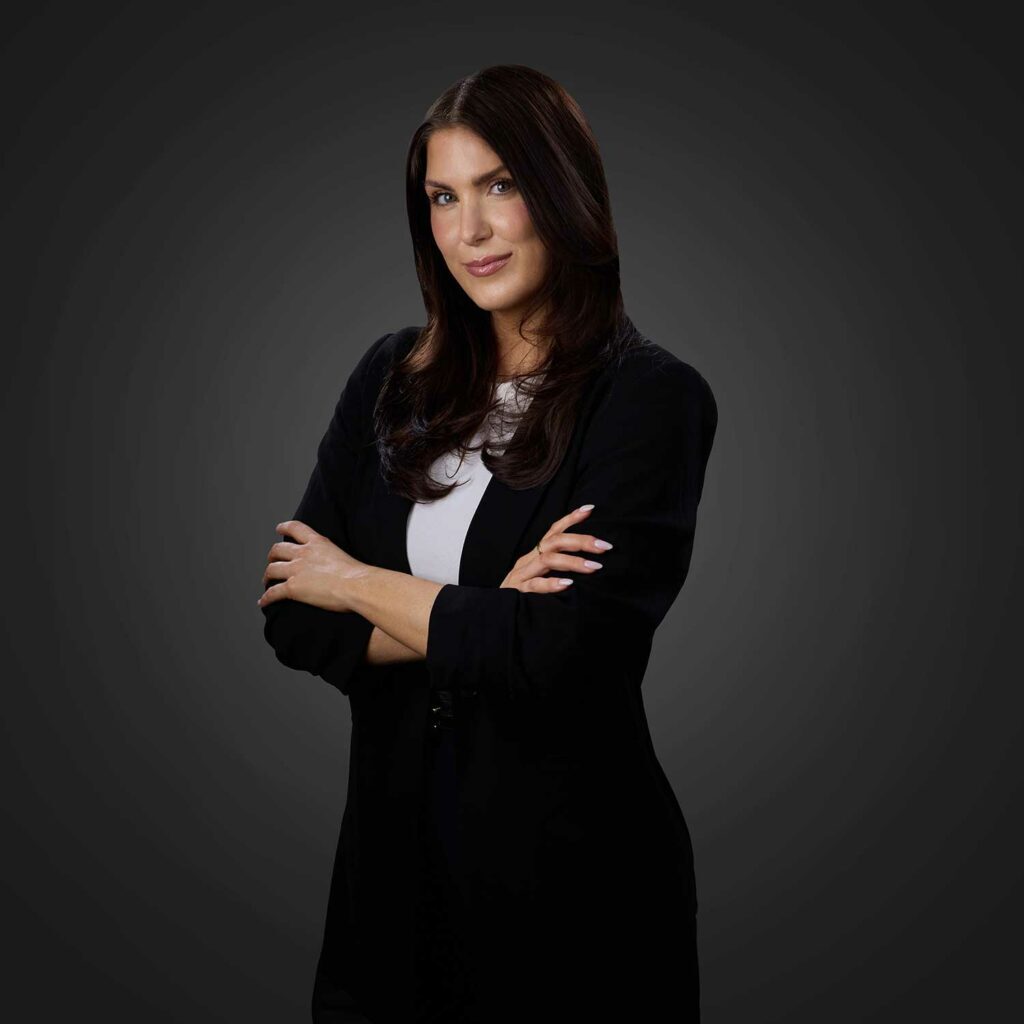 Portrait of a confident brunette woman with long hair, wearing a black jacket over a white top, standing with her arms crossed against a dark gradient background.