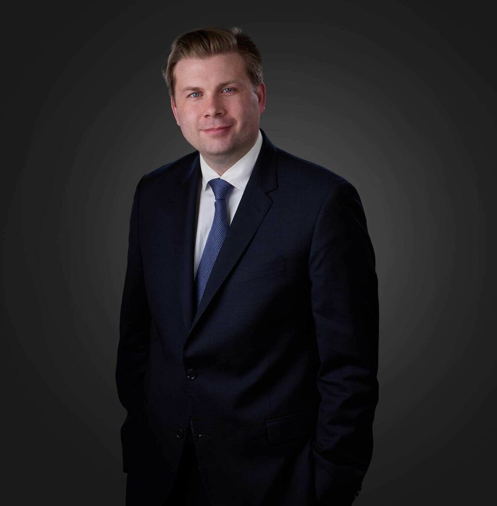 Portrait of a man with light brown hair wearing a dark suit, white shirt, and blue tie, standing against a dark gradient background with hands in pockets and a slight smile