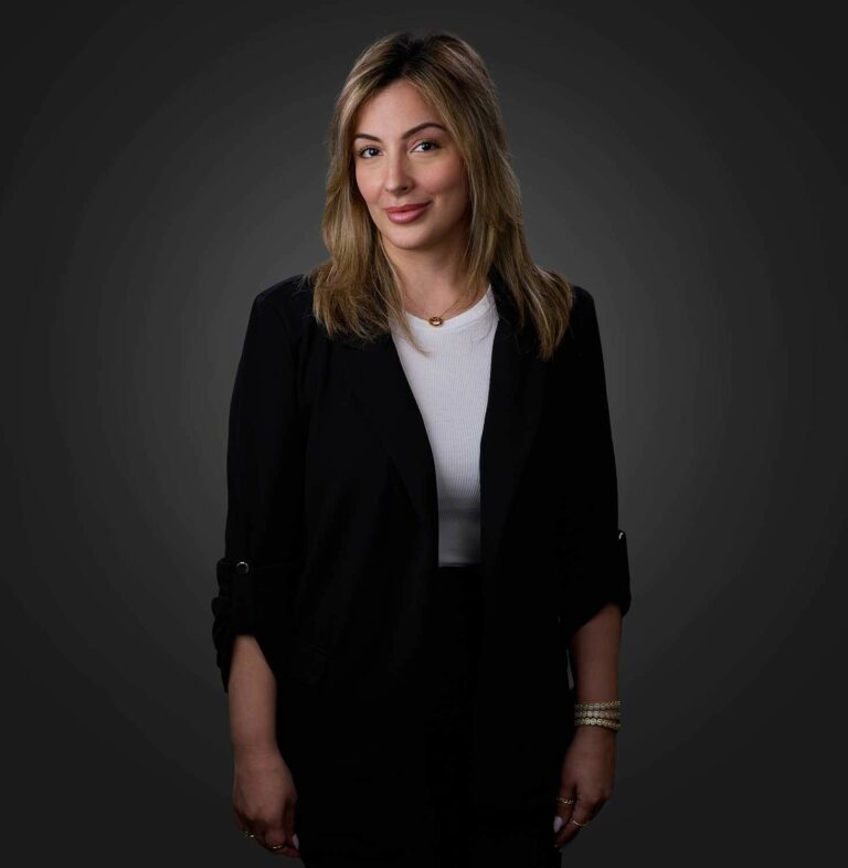 Smiling woman with light brown hair wearing a black blazer and white top, standing against a dark gray background