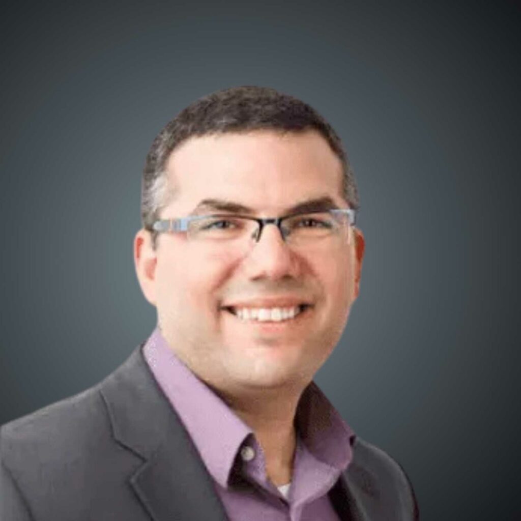 Portrait of a smiling man with short dark hair and glasses, wearing a dark blazer and a purple collared shirt, against a plain dark background.