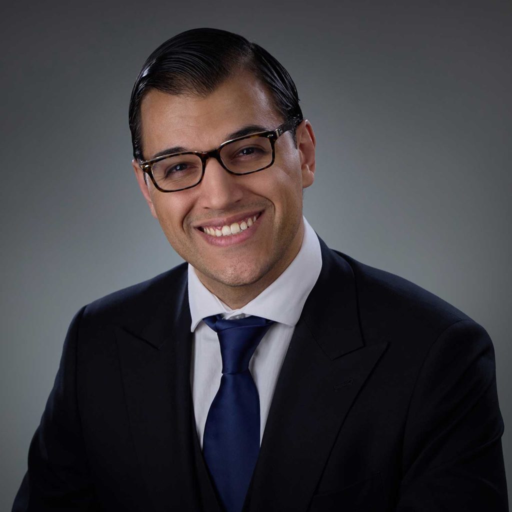 Man with neatly combed dark hair wearing tortoiseshell glasses, a dark suit, white shirt, and blue tie, smiling against a neutral gray background