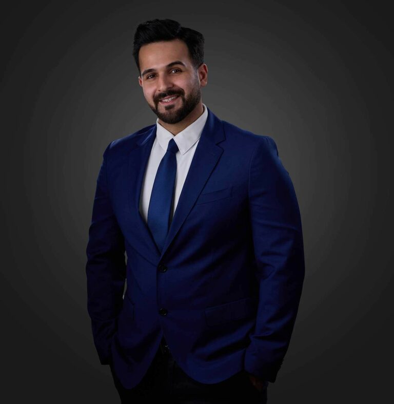 Smiling man with beard wearing a dark blue suit, white shirt, and blue tie, standing against a dark gray background.