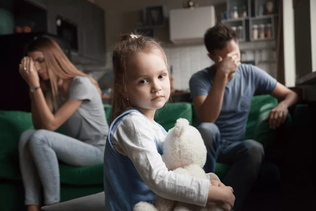 Young girl holding a white teddy bear looking sad in the foreground with distressed parents sitting apart on a couch in a home setting, representing family conflict and the need for family law support in Toronto.