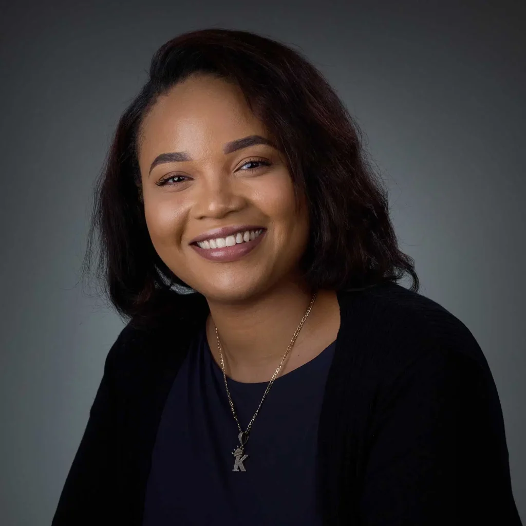 Smiling woman with shoulder-length dark hair wearing a black top and a gold necklace with a 'K' pendant against a gray background.