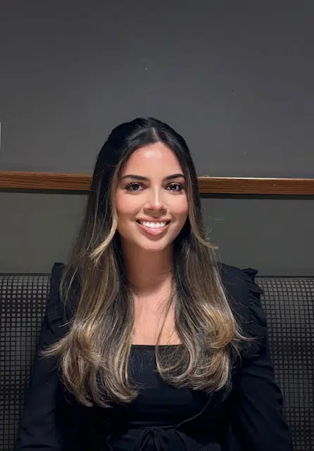 A young woman with long, dark brown hair with blonde highlights, wearing a black top, smiling and seated against a dark background with a wooden trim.