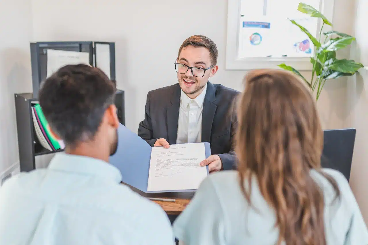 A family lawyer in Toronto from Nussbaum Family Lawyers wearing glasses and a suit, explaining legal documents to a couple in a bright office setting with certificates on the wall and a plant nearby.