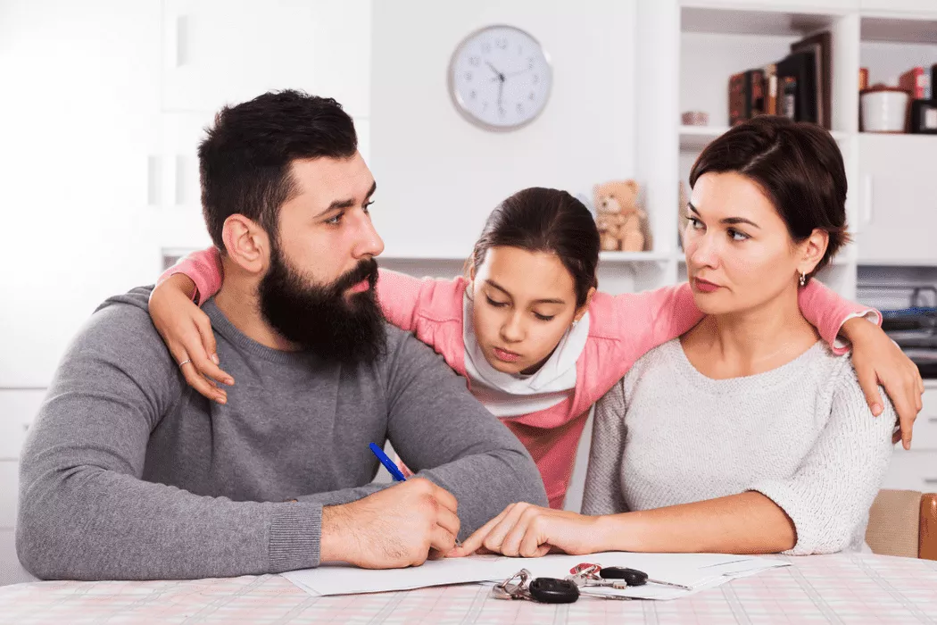 A serious family of three, with a man writing on a document at a table, a woman looking concerned, and a child with arms around both parents, symbolizing a family consultation possibly with Nussbaum family lawyers in Toronto.