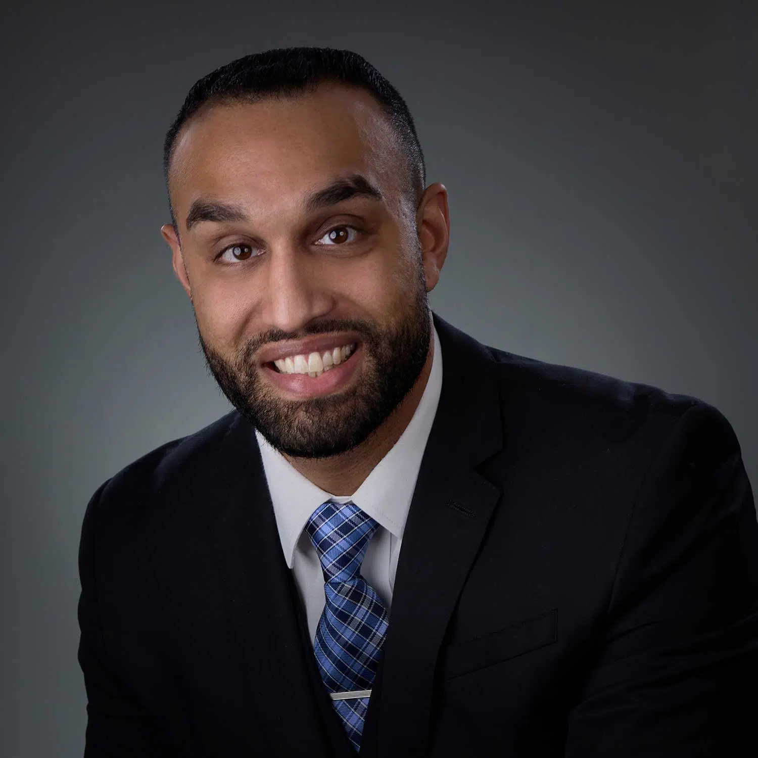 Man with short dark hair and beard, smiling, wearing a black suit jacket, white dress shirt, and blue plaid tie, against a plain gray backdrop.