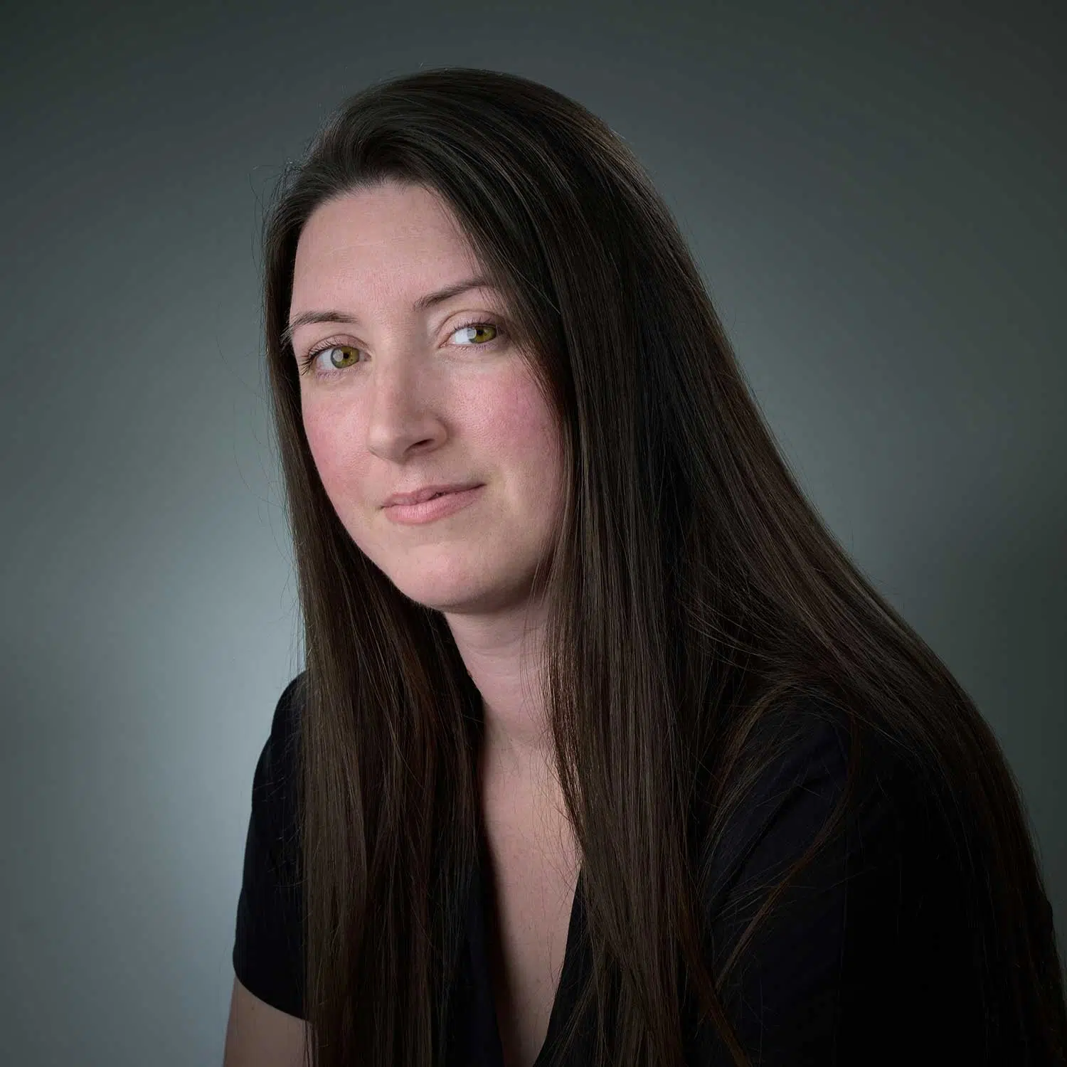 Portrait of a woman with long dark brown hair and light eyes, wearing a black top, against a neutral gray background.