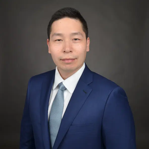 Portrait of an Asian man wearing a blue suit jacket, white shirt, and light blue tie, standing against a plain dark background.