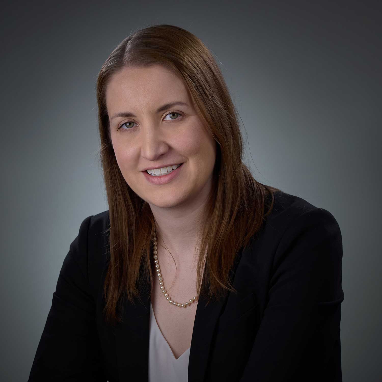 Smiling woman with straight brown hair wearing a black blazer, light blouse, and a pearl necklace against a gray gradient background