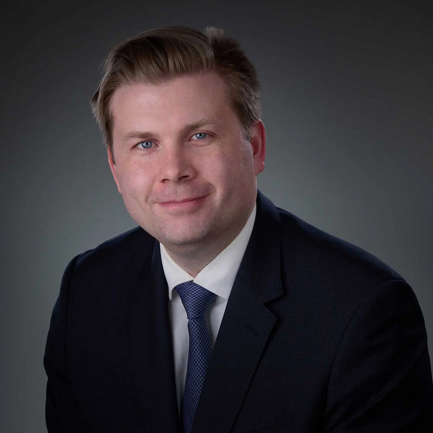 Portrait of a man with light brown hair wearing a dark suit, white shirt, and blue tie against a gray gradient background.