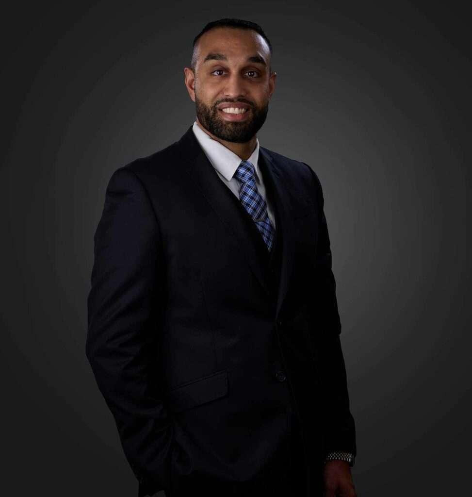 Smiling man with a beard wearing a black suit, white shirt, and blue checkered tie, standing against a dark gray gradient background with one hand in his pocket.