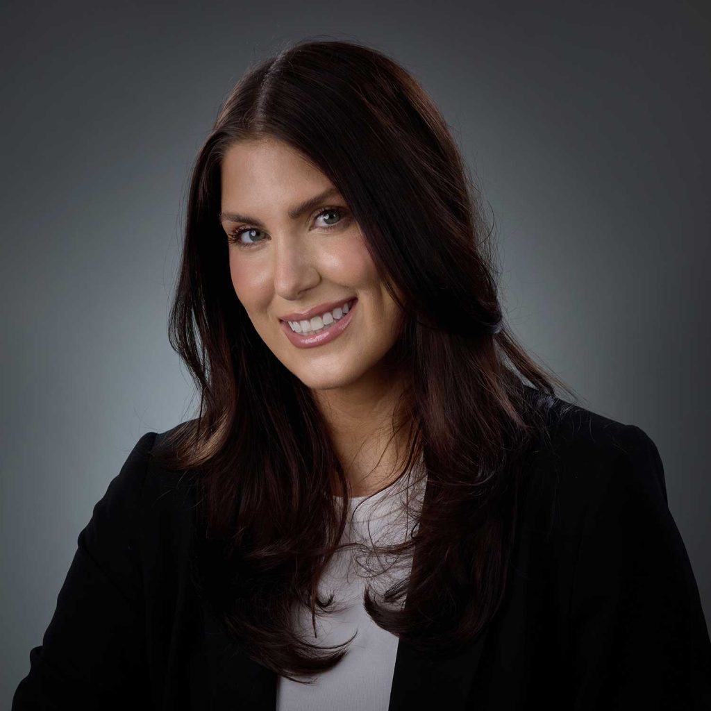 Smiling woman with long dark brown hair wearing a black blazer and white top against a gray background.