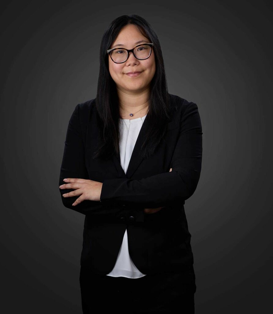 Confident woman with long black hair and glasses wearing a black blazer over a white blouse, standing with arms crossed against a dark gray background