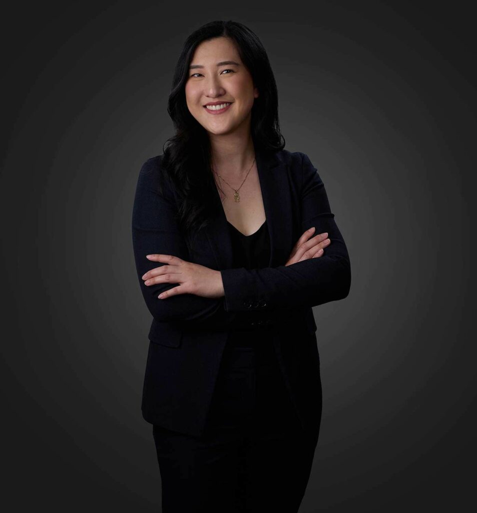 Smiling woman with long black hair wearing a black blazer and top, standing with arms crossed against a dark gradient background.