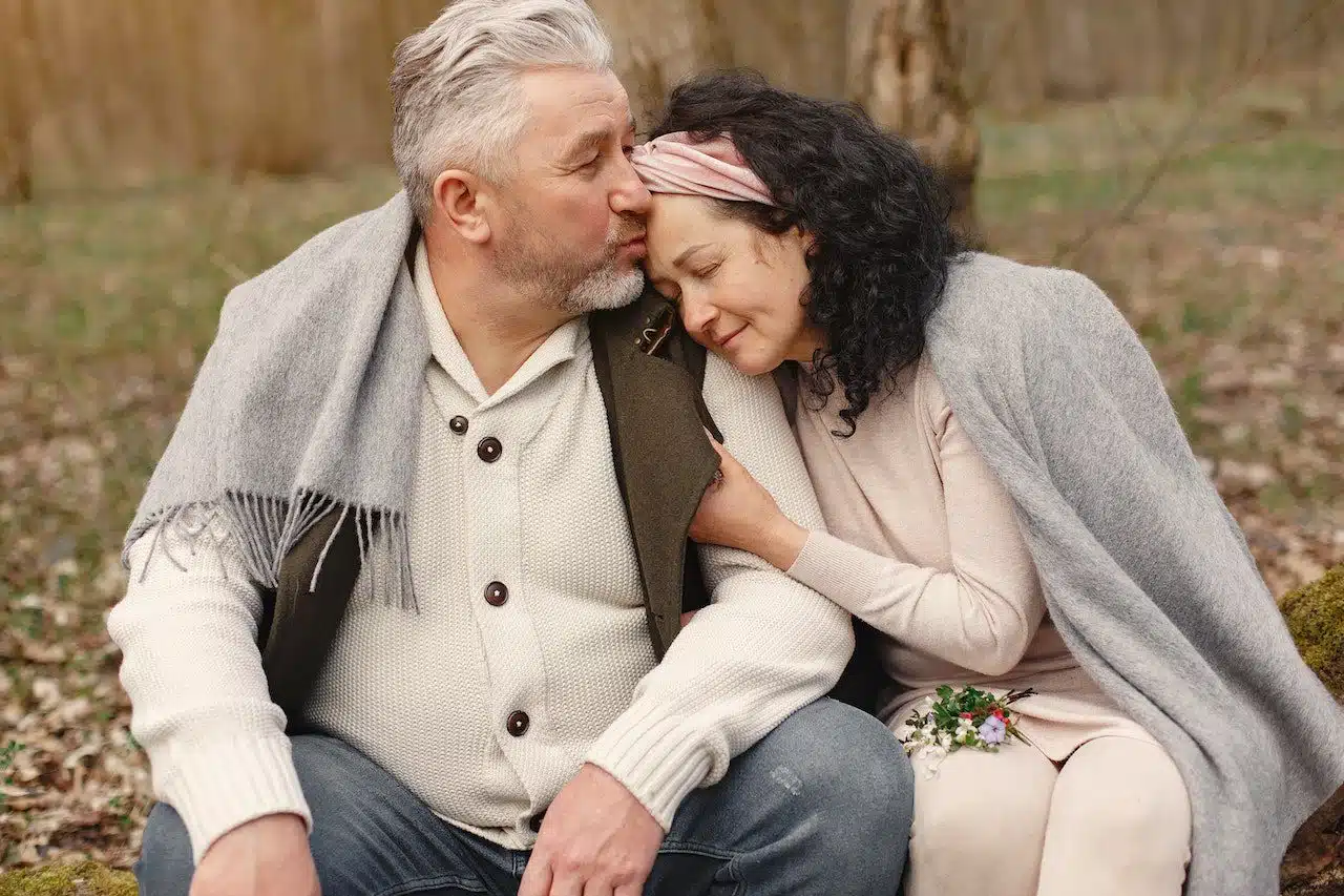 An older couple sitting close together outdoors wrapped in a gray blanket, sharing a tender moment with the man gently kissing the woman's forehead, symbolizing supportive care and trust relevant to family lawyers in Toronto.