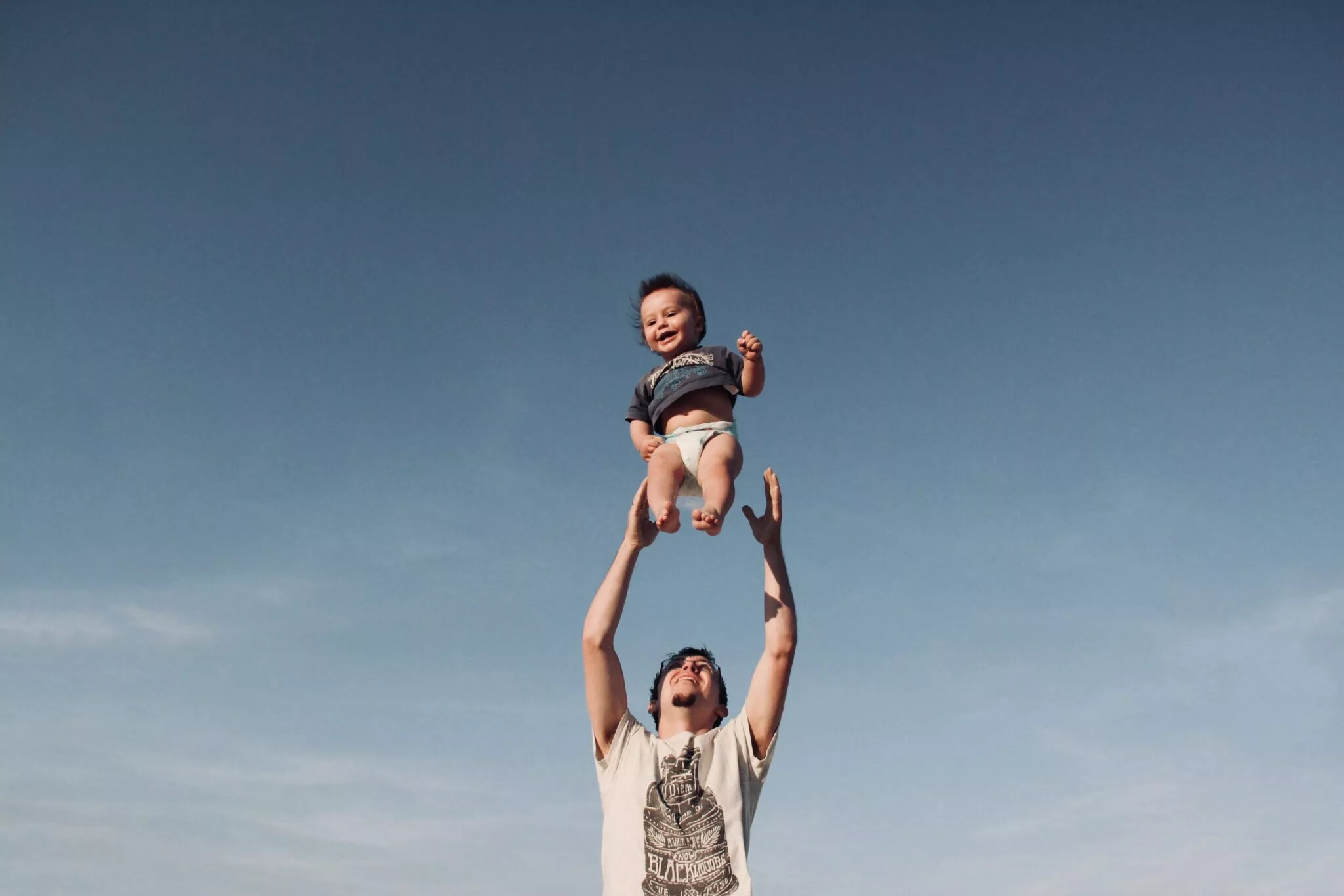 A smiling father joyfully tossing his baby into the air under a clear blue sky, symbolizing family care and trust, relevant to Nussbaum family lawyers in Toronto