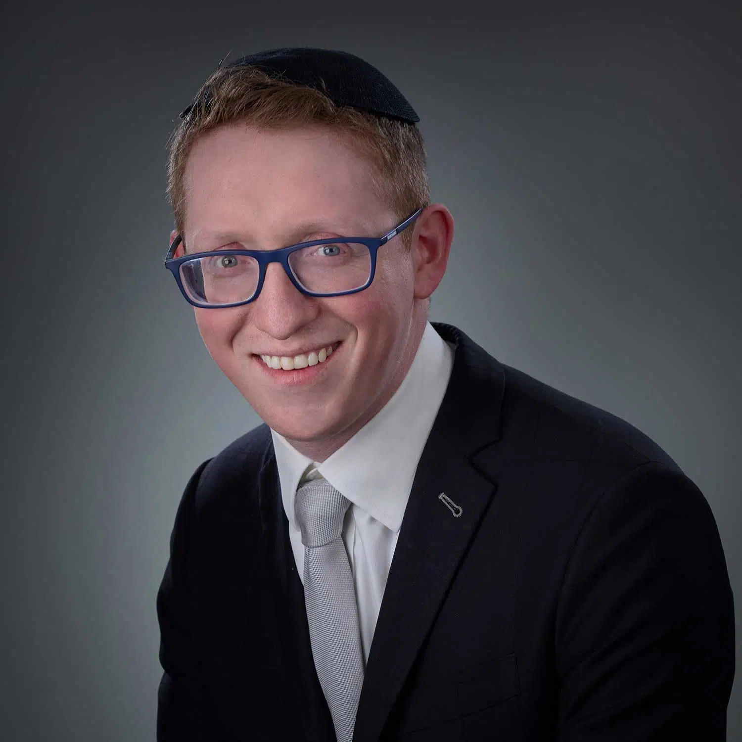 Smiling man wearing blue glasses, a black kippah, a dark suit, white shirt, and light gray tie against a gradient gray background.