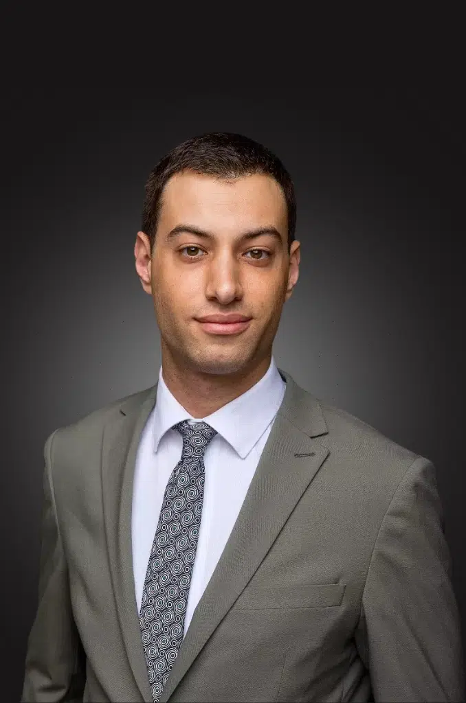 Portrait of a young man in a light gray suit, white shirt, and patterned tie against a dark background.