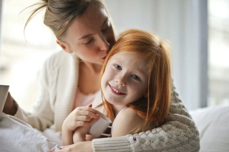 A loving mother gently kissing and hugging her smiling young daughter indoors, symbolizing family care and support relevant to Nussbaum family lawyers in Toronto.